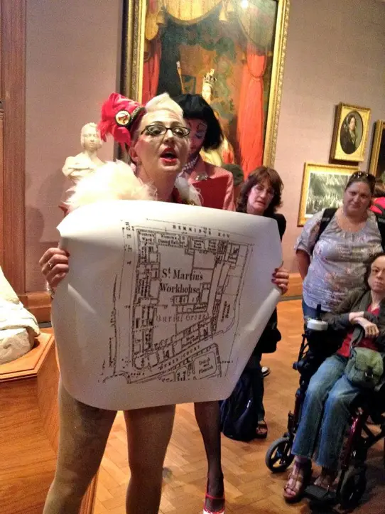 A photo from one of Bird's performances at the National Portrait Gallery. Bird is holding up a ground plan of St Martin's Workhouse. The audience is standing around her.