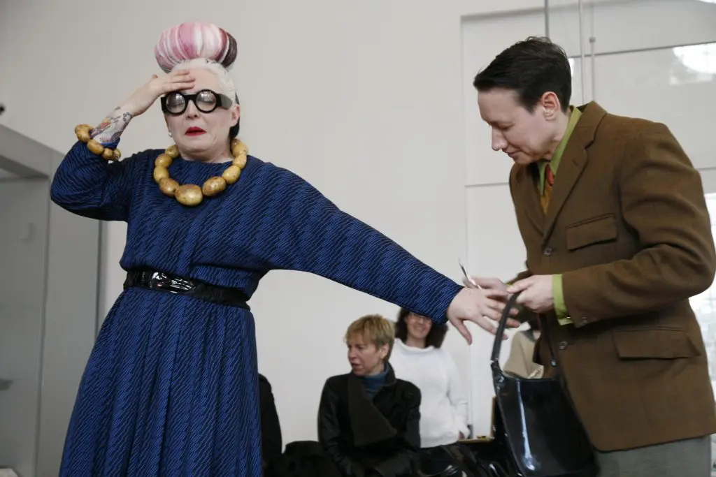 Birdie is gesturing towards her head while her handsome besuited butch husband passes her some pills from her handbag.