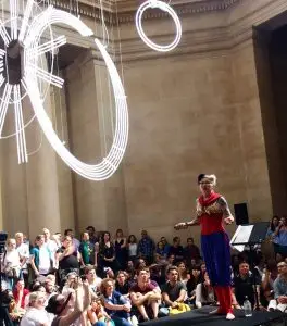 Bird holding forth on a podium in a large space in Tate Britain. She is wearing her Queer Communards red and blue outfit and is surrounded by the audience.